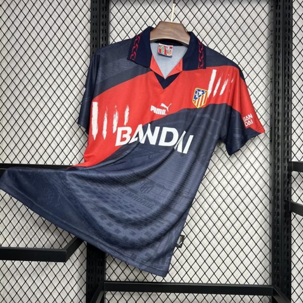 Camiseta visitante Atlético de Madrid 1996-97 en azul marino con franja diagonal roja y blanca, escudo bordado y patrocinio Bandai